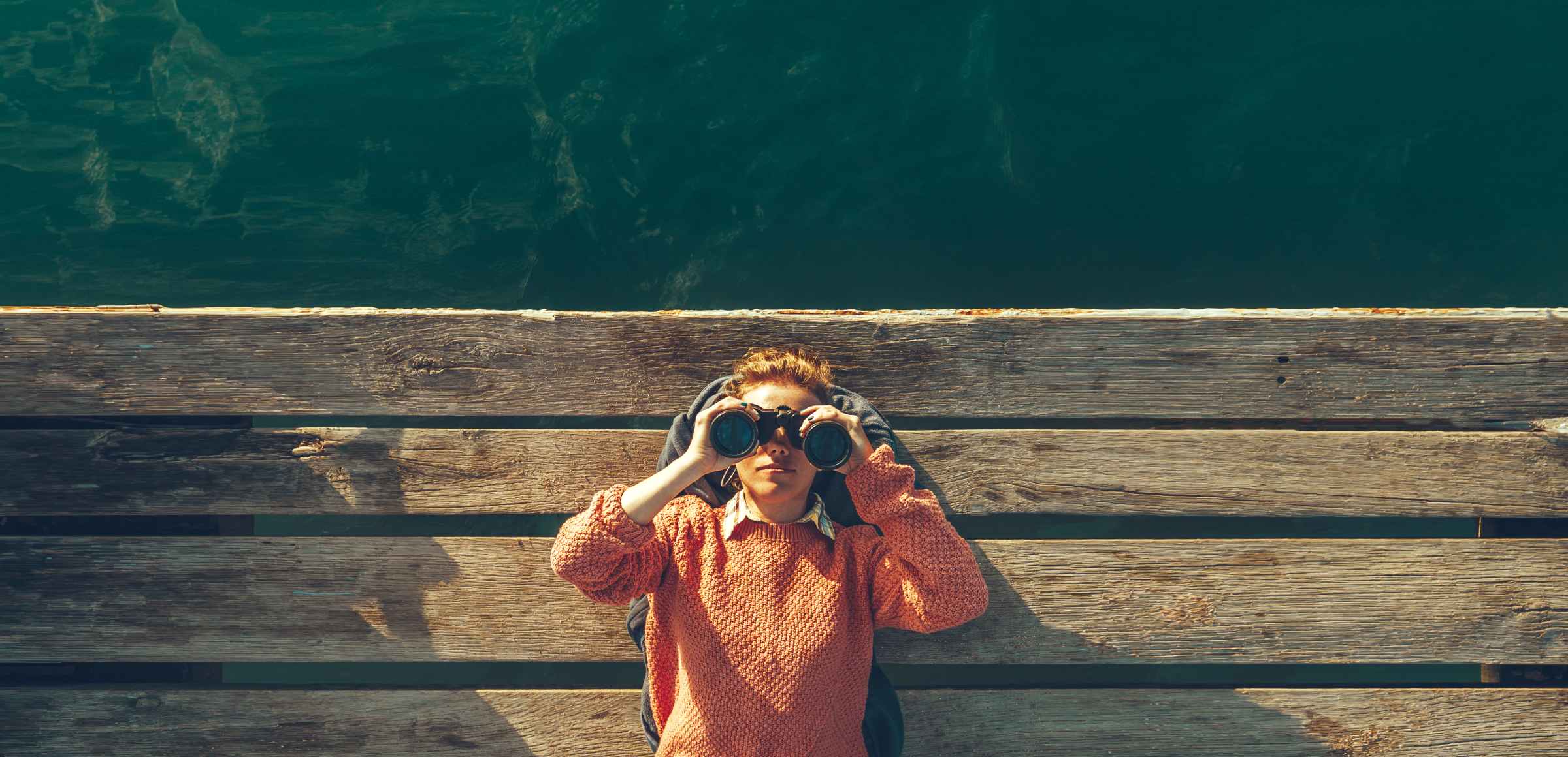 Ragazza sdraiata su un molo in riva al mare guarda attraverso un binocolo.  Concetto di curiosità
