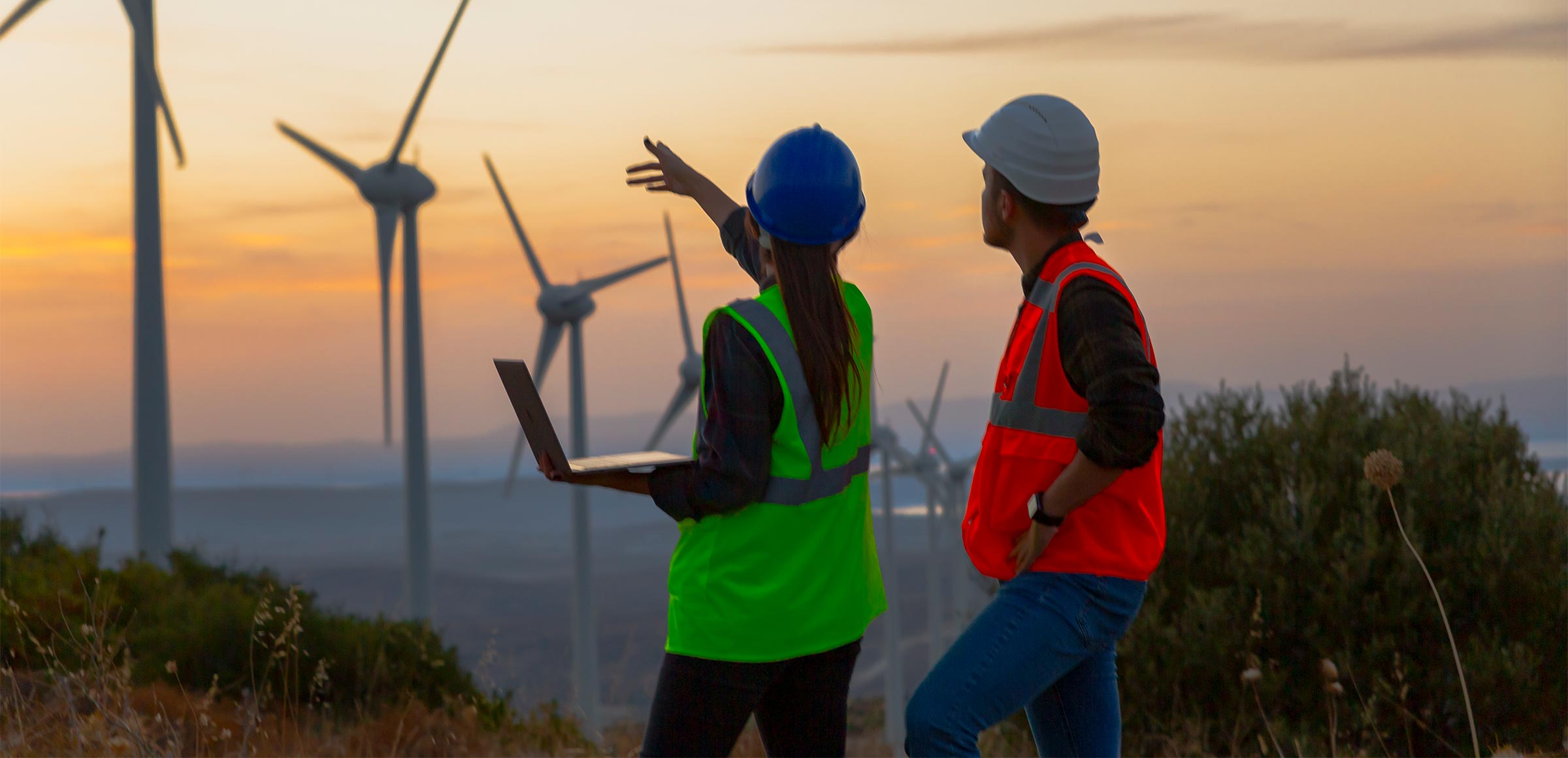 Young electrical engineer woman and business man standing in front of a wind turbine park at sunset